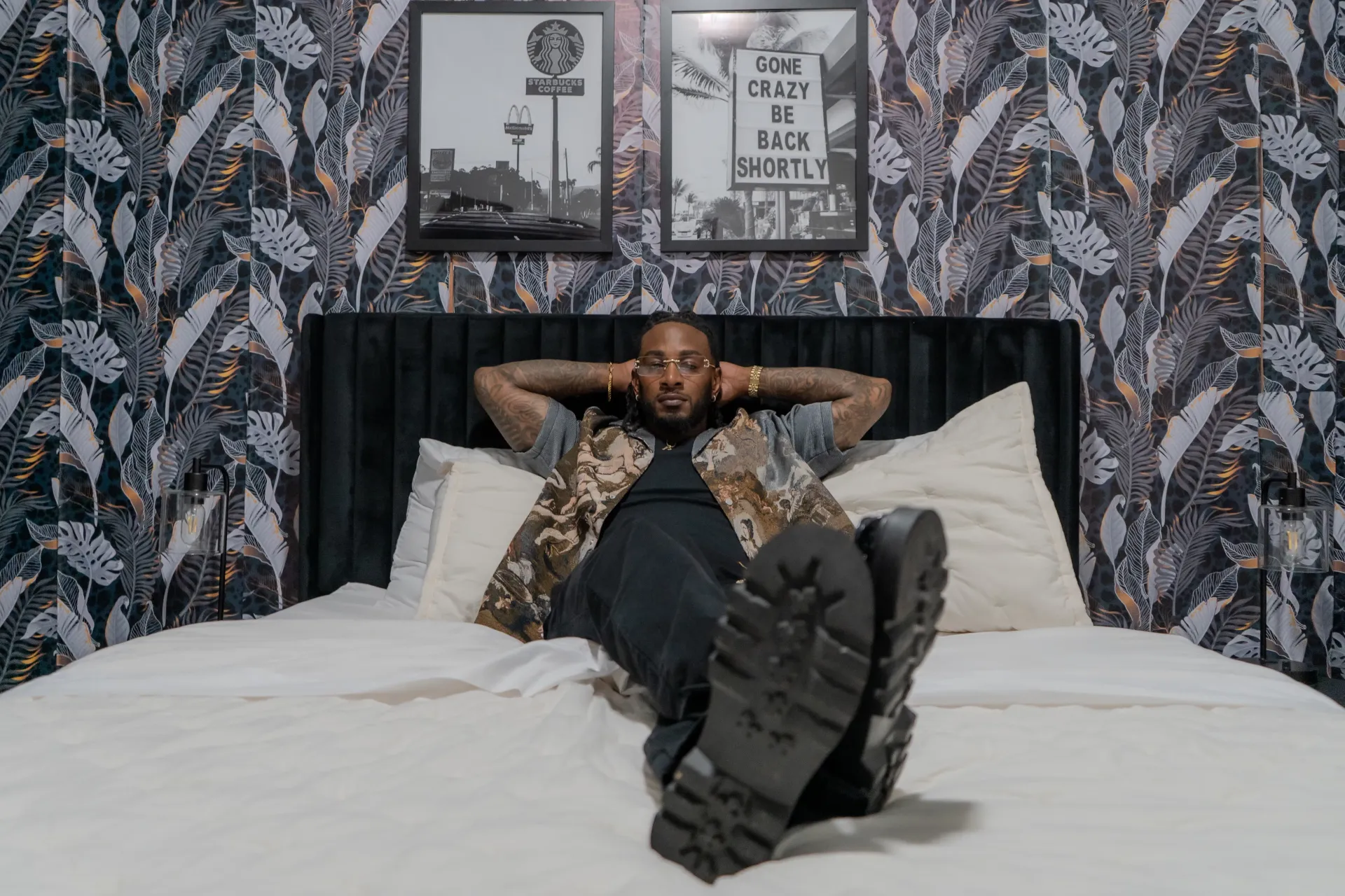 Brian Humble Hampton relaxing on a bed in a camouflage shirt and gold-rimmed glasses